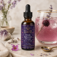 An amber glass dropper bottle labeled 'Oceanside Apothecary Drink Infusion Drops' stands on a marble surface beside a pink beverage in a glass, lavender sprigs, a spoon with dried lavender, and soft floral decor in a warm styled setting.
