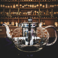 Clear glass teapot with a metallic lid against a colorful geometric patterned background