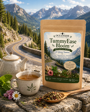 Load image into Gallery viewer, A stand-up pouch of Oceanside Apothecary TummyEase Bloom herbal tea is displayed on a stone surface with a teapot, cup of tea, flowers, and a spoonful of loose tea in the foreground. Behind it is a winding mountain road and pine-covered hills under a blue sky.