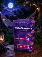 Load image into Gallery viewer, A bag of VitaDreamz Stumber Berry natural sleep aid gumdrops stands on a wooden table in a nighttime garden setting with a full moon, string lights, hammocks, and lavender. The packaging is purple with pink and white text, and several red-brown gummies are shown on the front.